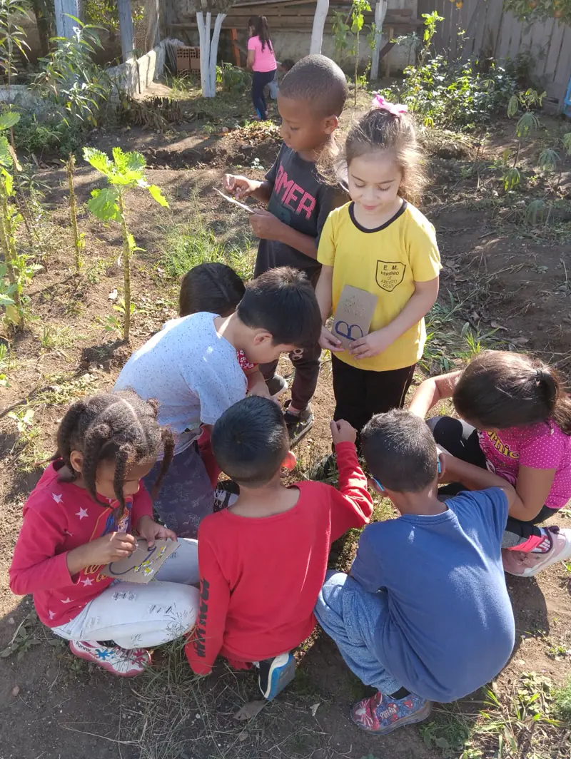 Crianças agachadas plantando mudas na terra da horta comunitária